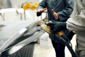 A person speaking with an auto repair technician while standing by a car with visible damage in a repair shop.